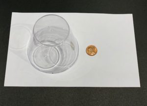 Materials needed for magic trick, including an upside down clear plastic cup next to a penny.