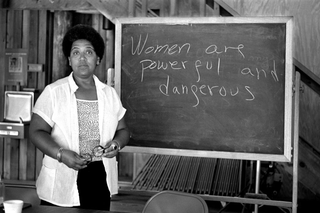 Black and white photo of Audre Lorde in front of a chalk board that reads "Women are powerful and dangerous."