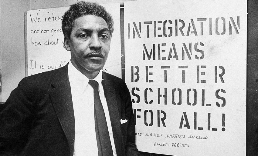 Black and white photo of Bayard Rustin in front of a sign that reads "Integration means better schools for all!."