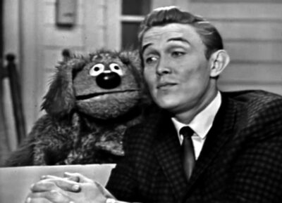 black and white still from the Jimmy Dean Show depicting Dean with a dog puppet, Rowlf, peering over his shoulder