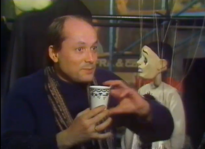 grainy video still depicting a man holding a cup in two hands, a marionette hanging behind him