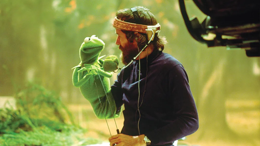 green-tinged photo depicting a man, Jim Henson, with a Kermit puppet on his arm, holding poles to control the puppet's arms, and wearing a headset for voicing the puppet