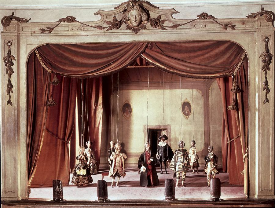 photo depicting several small marionettes strung up in a theater with red curtains