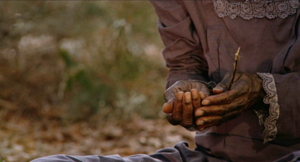 film still depicting a woman's torso sitting in front of blurred grass. She wears a dull purple dress and holds out her hands, which have dark indigo stains across them