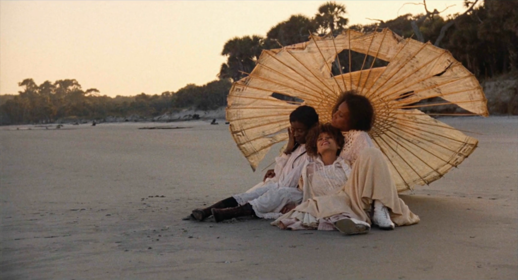 film still depicting three women lounging on a beach under a tattered worn umbrella