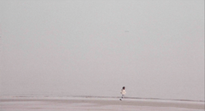 film depicting a small speck of a toddler running across a wide frame of a misty beach with white ocean filling the frame