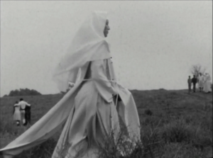 black and white film still depicting a nun in habit walking across the grass in front of two groups of people in the distance