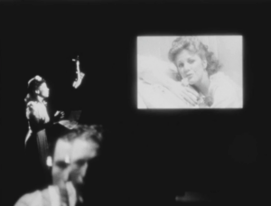 Black and white film still depicting a woman singing into a mic while a film plays on a screen across from her. Reflection of sound engineer is seen in the glass