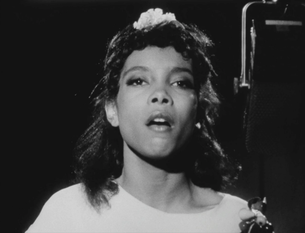 black and white film still depicting a young woman singing, her face fills the frame