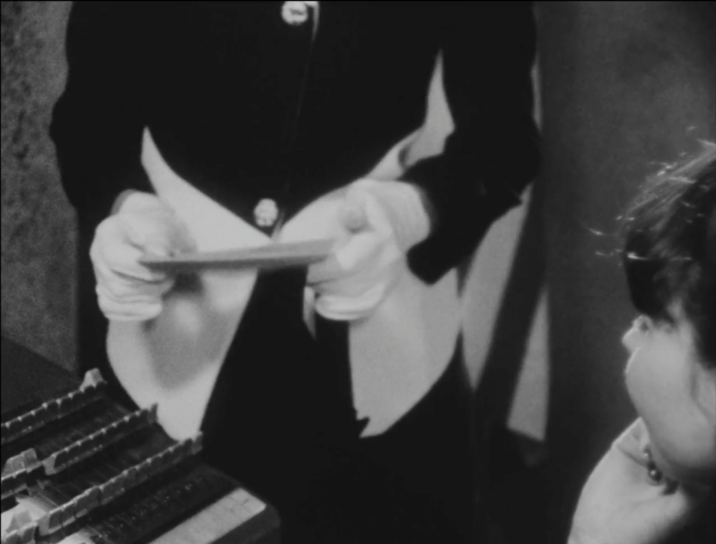black and white film still depicting the lower half of a woman in a pant suit holding a letter while a the profile of a second woman's face is shown looking up at the first