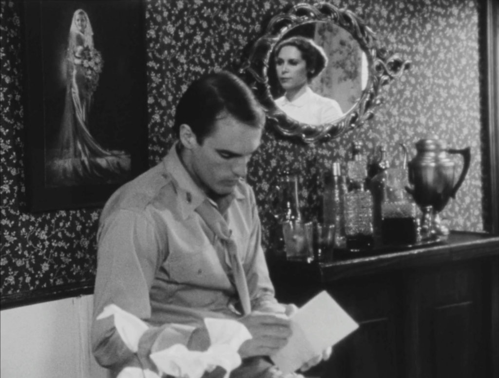 black and white film still depicting a man seated at a desk while a woman speaks to him offscreen, her face reflected in the mirror behind him