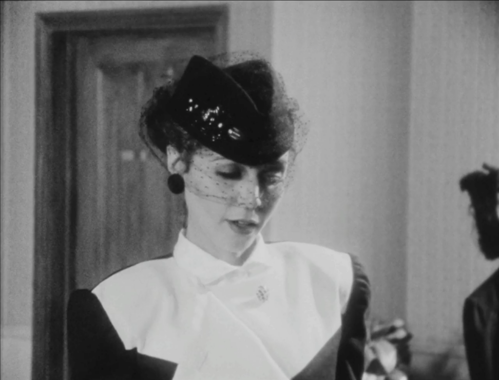 black and white film still depicting a woman from the shoulders up wearing a black hat with a black dot chenille veil obscuring her face