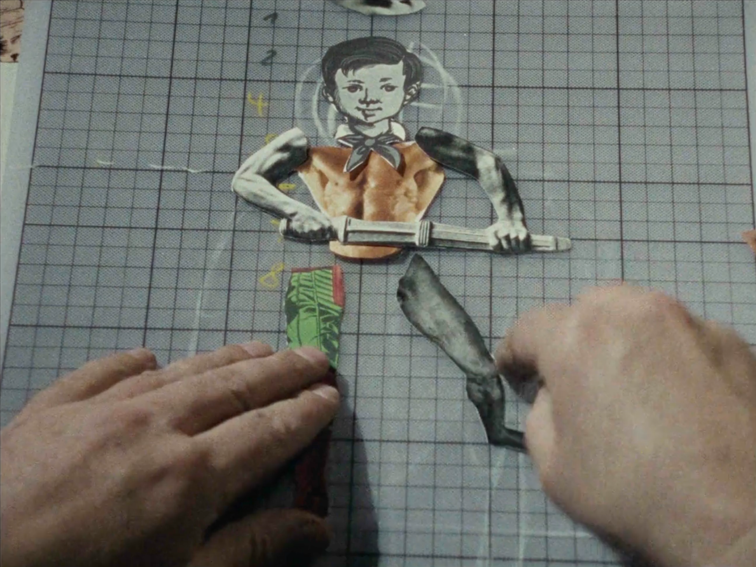 film still depicting a man's hands holding paper cut-out legs as he pieces together a humanoid figure on top of a gray grid cutting mat
