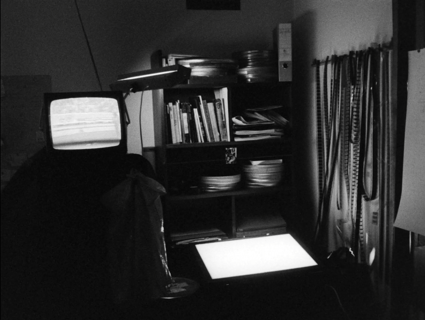 black and white film still depicting the corner of an artist's studio with film reels and books filed on a shelf, loose film hanging on the wall, a light table and a fuzzy CRT TV
