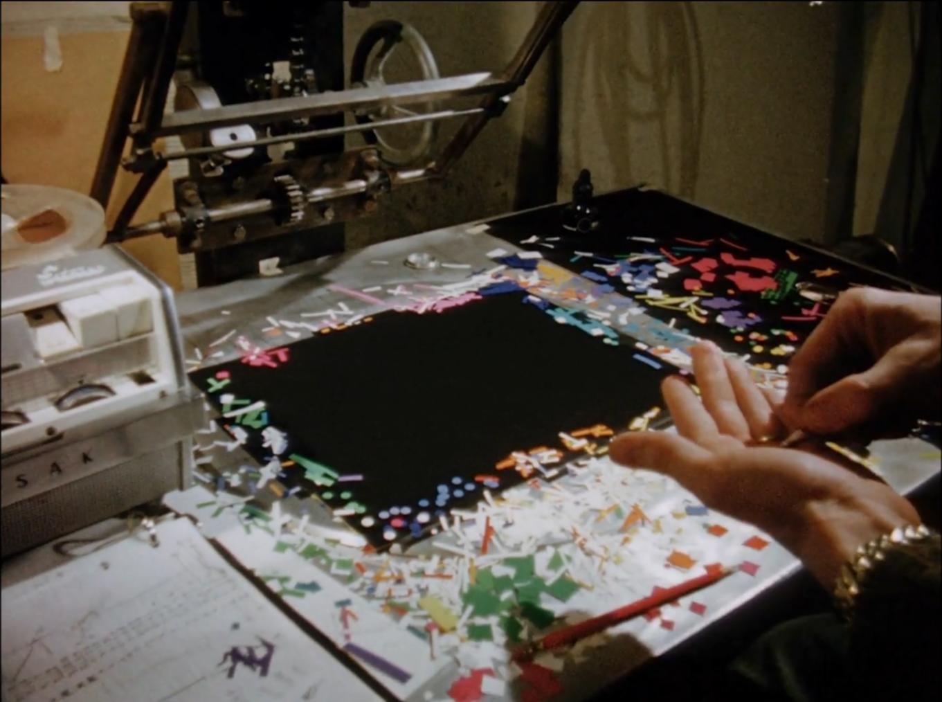 film still depicting a worktable used for stop-motion animation with scraps of paper strewn across and a man's hands delicately places the pieces
