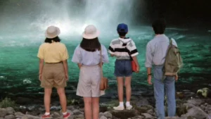 film still depicting four children standing center frame, gazing at a vibrant teal-colored pond at the bottom of a misty waterfall