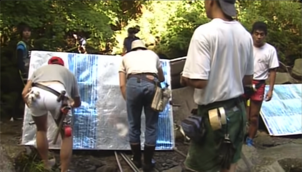 photograph of several men wearing utility belts, backs turned to camera, two bent over drawing blue lines onto a shiny silver light reflector