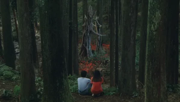 film still from Rhapsody in August depicting a dark, moody forest with two children kneeling center frame, looking out at a clearing full of red flower and a dead tree