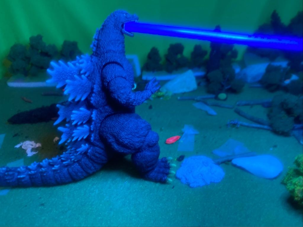 film still depicting a Godzilla toy firing a blast out of its mouth while the scene is bathed in UV light