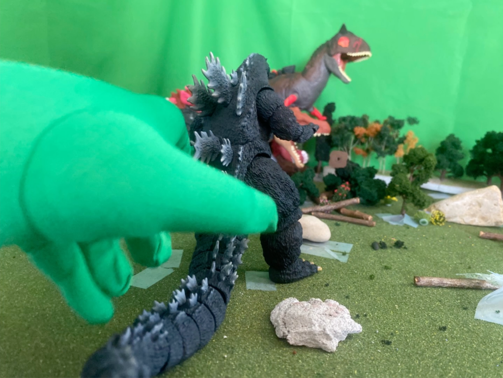 film still depicting a scene of plastic monster toys fighting while a green gloved hand reaches in to pose on of the figures