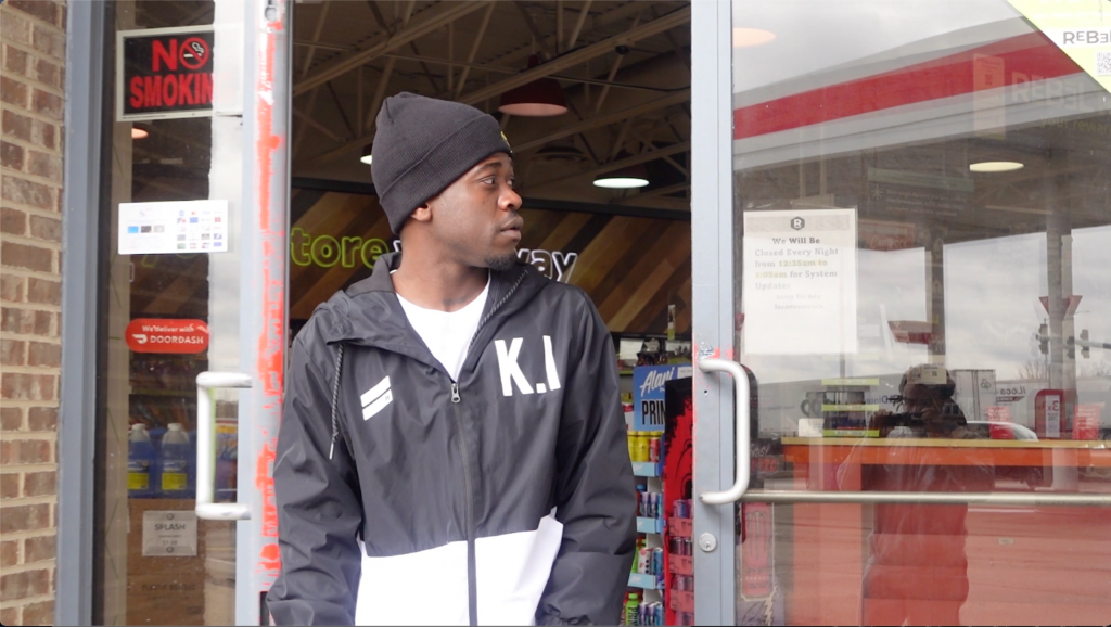 film still depicting a young man stepping out of the glass-lined threshold of a gas station
