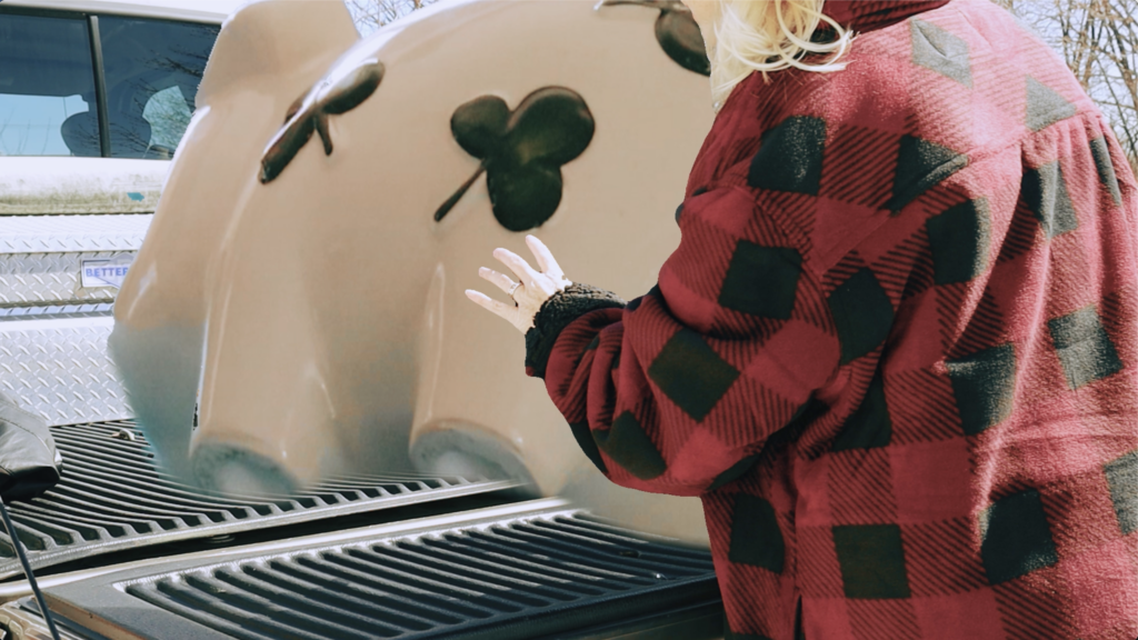 film still depicting the torso of woman in plaid with arms extended as she pushes a giant pig into the back of a pickup truck