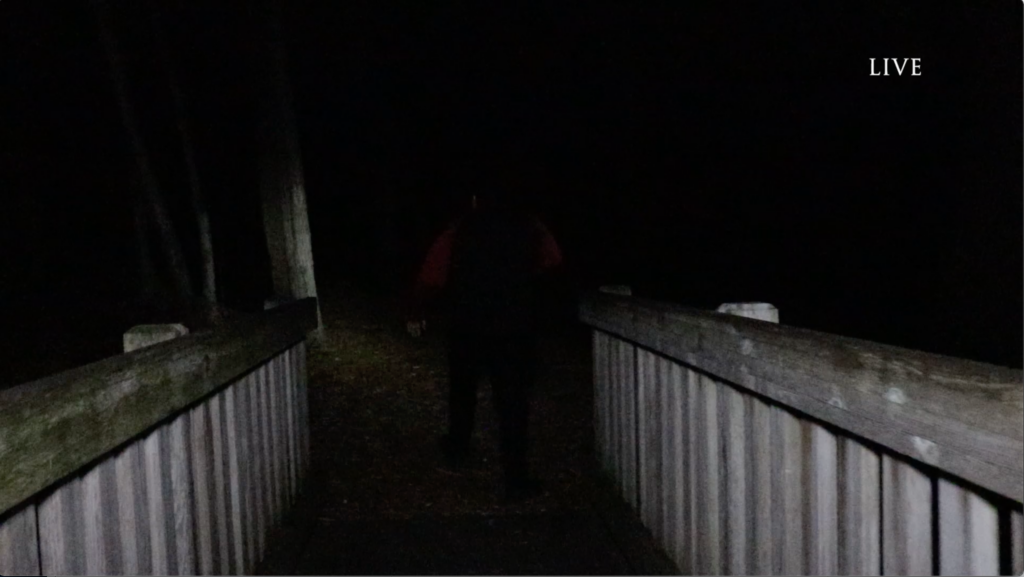 film still of a scene bathed in darkness, just a littler light spills onto the rails of a bridge as a man disappears into the darkness ahead. A small LIVE graphic sits in the upper right corner