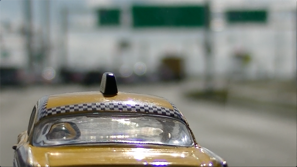 film still showing a very shiny yellow taxi driving toward a blurred highway signs in the distance
