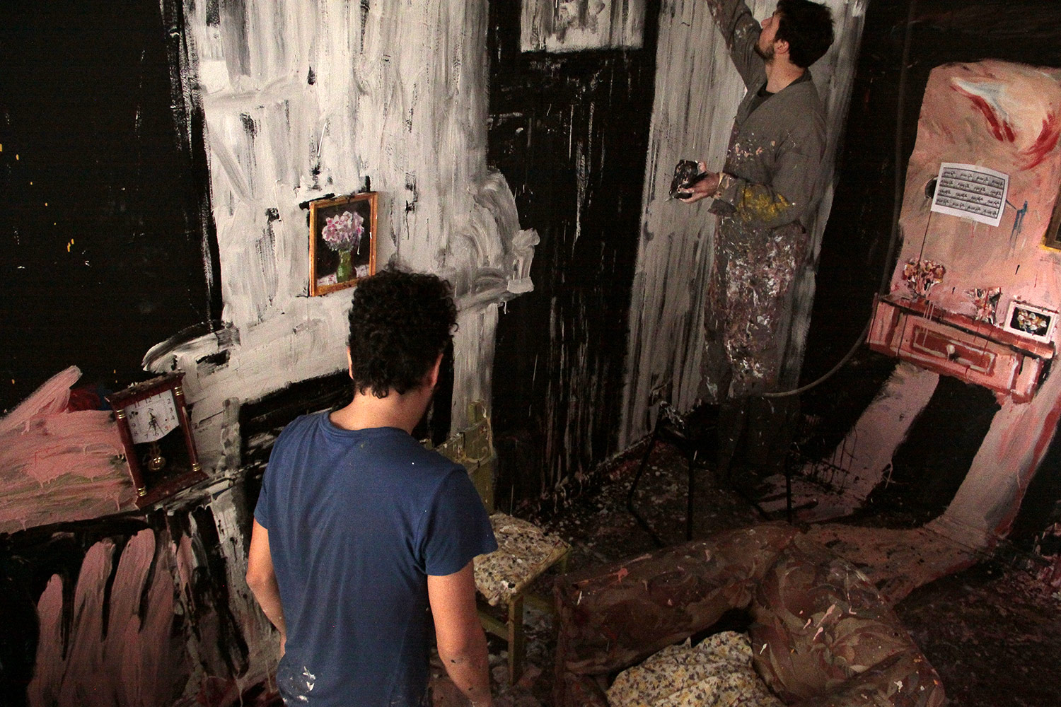 photograph depicting a messy studio space with one artist painting the wall and the other walking through the room