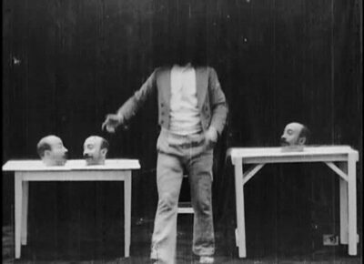 black and white film still depicting a headless man between two tables covered in disembodied heads