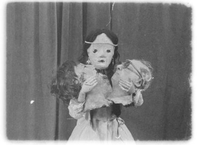 black and white film still depicting a young girl with twin braids holding two disembodied heads in front of a dark curtain
