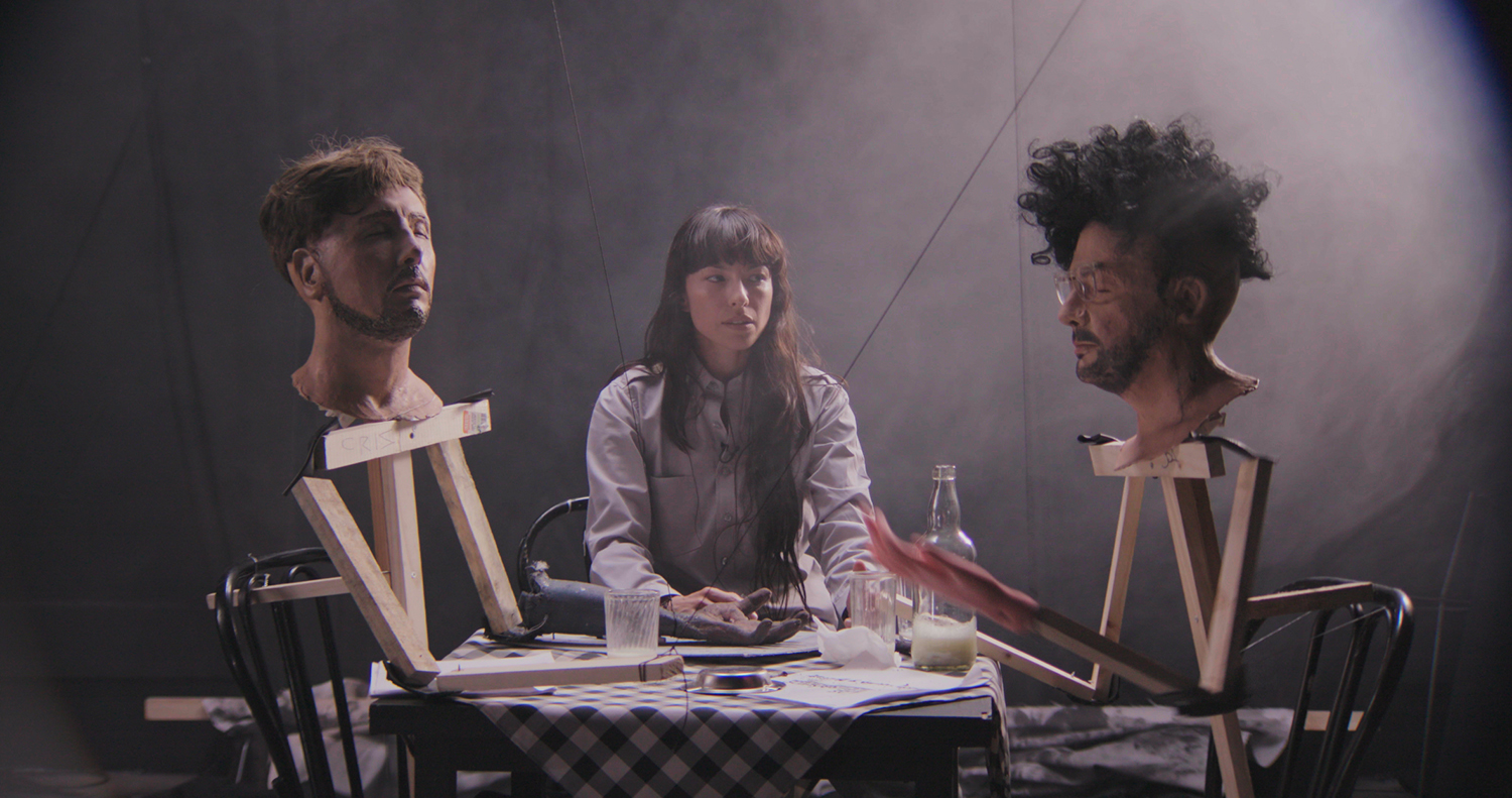 film still depicting a woman sitting at a table with sculpted heads of the directors on stiff, wooden armatures 