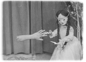 black and white film still of a puppet with twin braids reaching out to a disembodied hand