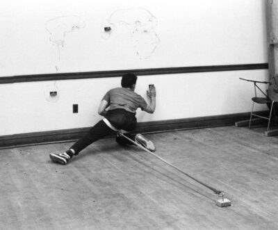 black and white photograph of a man seen from behind, kneeling awkwardly and drawing on the wall while a tether to the floor is tied around his thigh