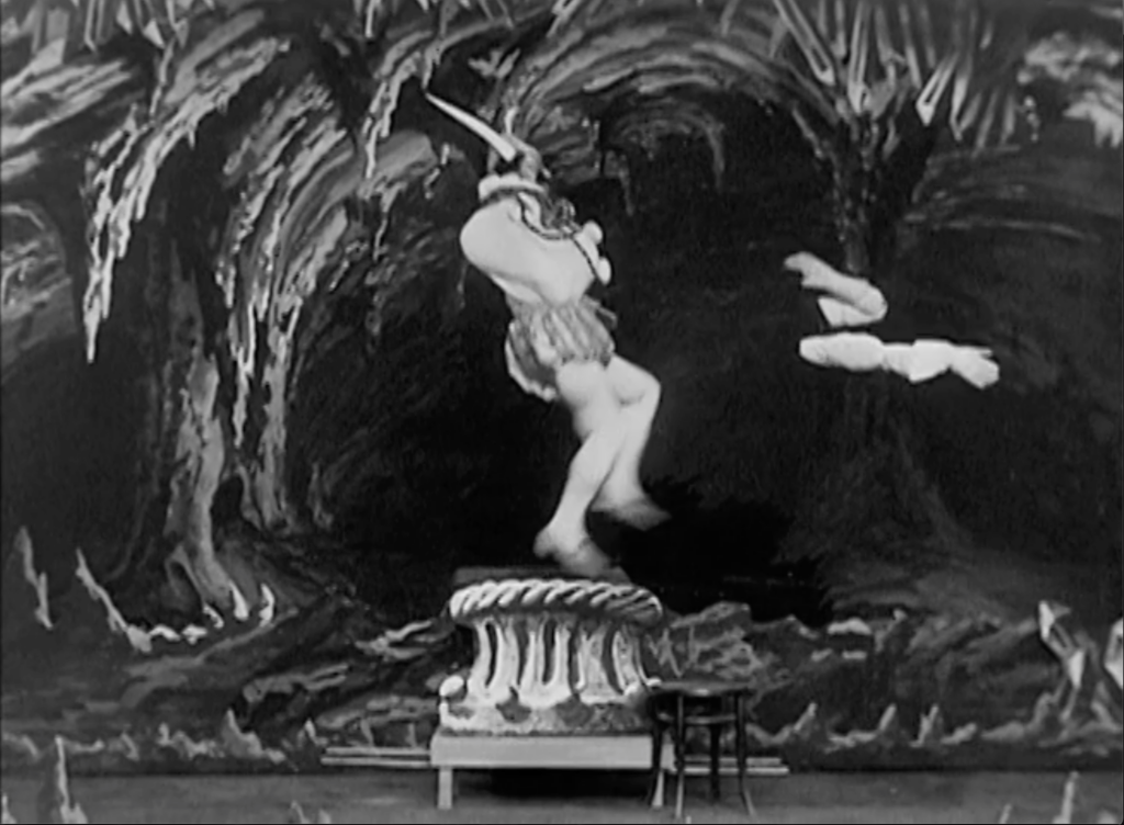 black and white film still of horned figure dancing on a cake in painted cave set, his arms disembodied to the right of the screen