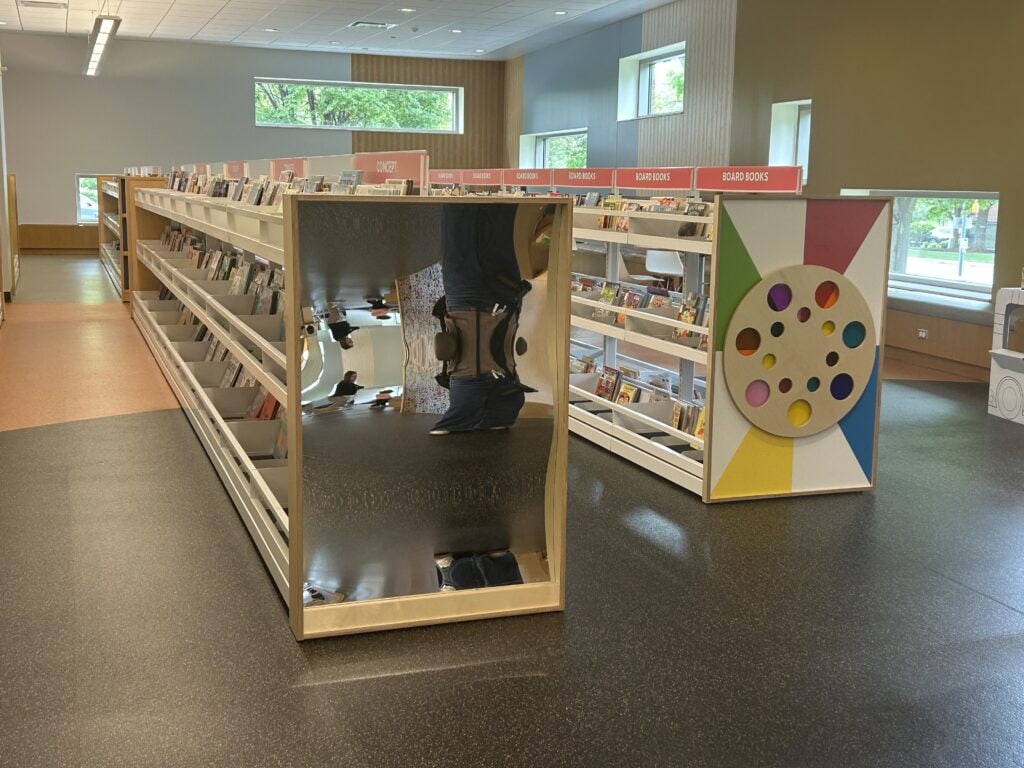 Children's library shelves with a large shiny mirror panel and a colorful circular wall feature.