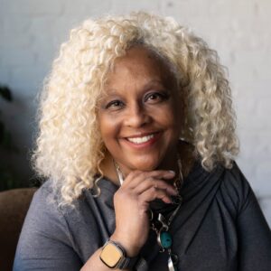 Woman with curly blonde hair sits in a brown chair, smiling at the camera. She wears a gray dress, necklace, smartwatch, and ring. White brick wall and plants are in the background.