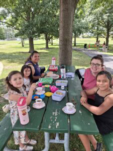 Two adults and four children sit at a green picnic table outdoors, having a meal with food, drinks, and dishes under the shade of trees in a park.