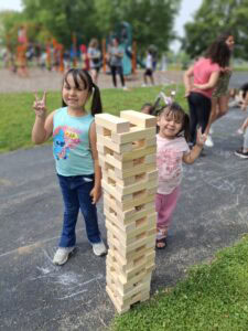 Two young girls stand beside a tall wooden block tower outdoors, both smiling and making peace signs; people and playground equipment are visible in the background.