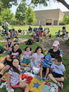 A group of children and adults sit on colorful blankets on the grass at an outdoor event, with trees and a building in the background.