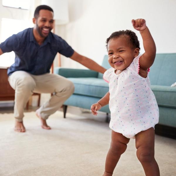 A smiling baby takes a step forward while an adult kneels and watches in the background in a living room.