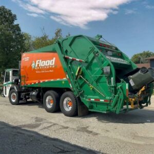 A green and orange Flood Brothers garbage truck is lifting and emptying a large black trash bin on a suburban street.