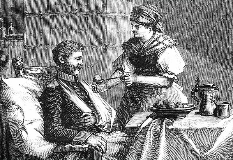 A woman feeds a seated man with an injured arm in a historical setting; food and drinks are set on a table beside them.