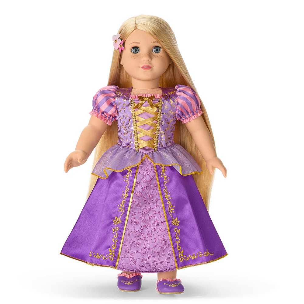 A blonde doll with long hair is wearing a purple and pink dress with gold trim and matching shoes, standing upright against a white background.