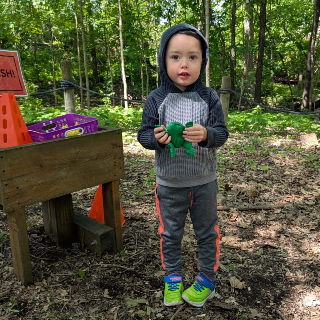 A young boy outdoors near an activity table, wearing a hoodie and holding a stuffed animal.