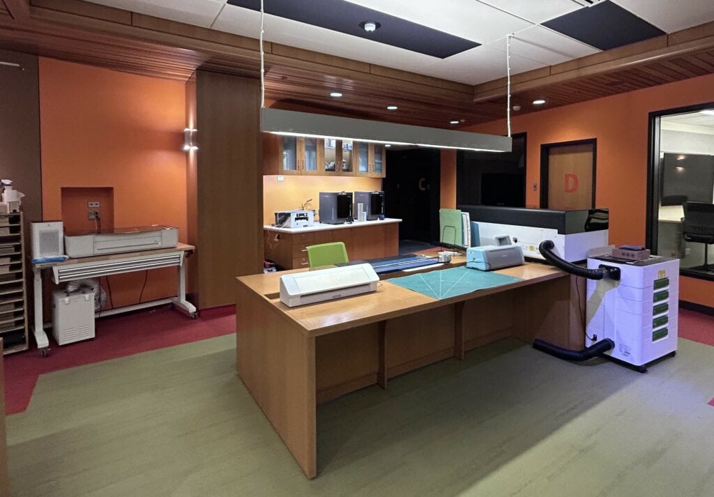 Office workspace with wooden desks, several printers, computers, and office equipment against orange walls, under ceiling lights.