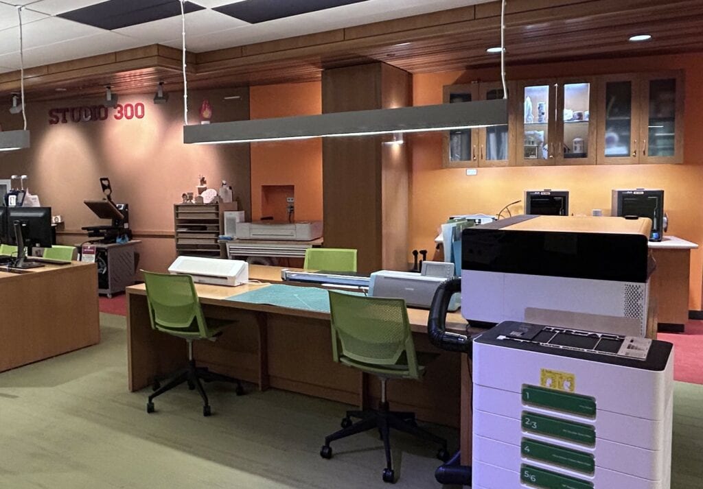 A workspace with several green chairs, equipment, printers, cabinets, and a sign reading “Studio 300” on an orange wall.