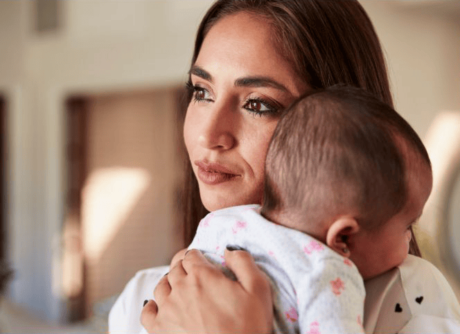 A woman holds a baby on her shoulder while looking into the distance, indoors with soft natural lighting.