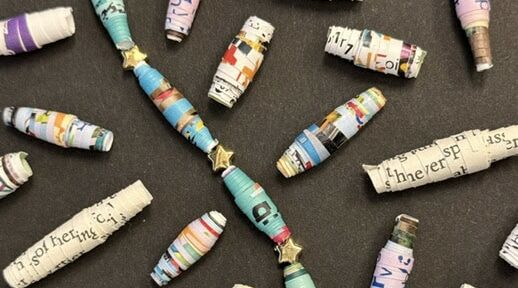 Colorful paper beads, some strung together and some loose, displayed on a dark surface. Each bead is made from rolled, printed paper strips.