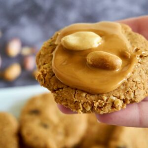 A hand holds an oat cookie topped with peanut butter and two whole peanuts, with more cookies blurred in the background.